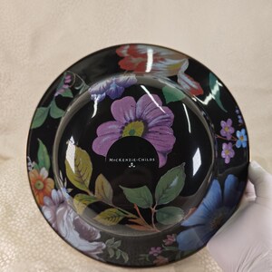 May include: A black decorative bowl with a floral design. The bowl has a glossy finish and is decorated with colourful flowers, butterflies, and green leaves. The text "McKENZIE-CHILDS" is visible on the bowl.