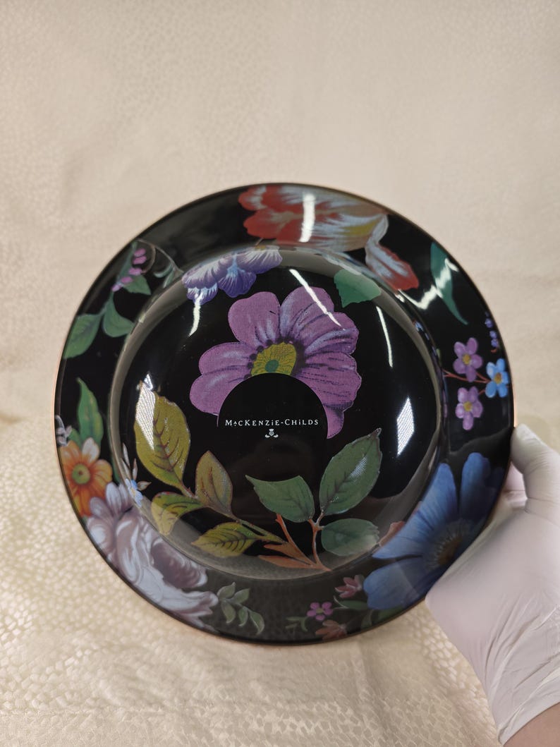 May include: A black decorative bowl with a floral design. The bowl has a glossy finish and is decorated with colourful flowers, butterflies, and green leaves. The text "McKENZIE-CHILDS" is visible on the bowl.