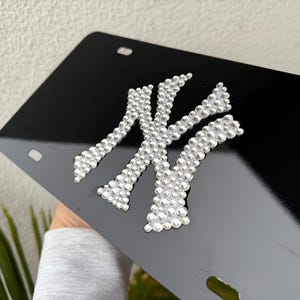 May include: A glossy black license plate frame featuring the New York Yankees logo rendered in clear, sparkling rhinestones. The rectangular plate has four mounting holes and displays the iconic interlocking NY emblem centered on the surface.