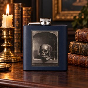 May include: A navy blue rectangular metal flask with a silver screw-top cap. The front features a centered rectangular print depicting a human skull resting on a book inside an arched alcove, accompanied by a small candle holder.