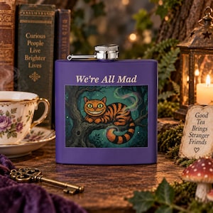 May include: A purple metal flask featuring a graphic of an orange striped cat with a wide grin perched on a tree branch. The text "We're All Mad" is printed in white above the illustration. The flask has a silver screw-top cap at the centre.