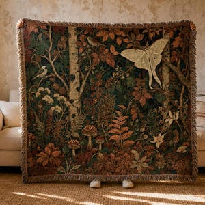 May include: A rectangular woven throw blanket featuring a detailed forest scene with a large cream-colored moth, various mushrooms, ferns, and trees in shades of deep green, brown, and rust orange, finished with a fringed edge on all four sides.