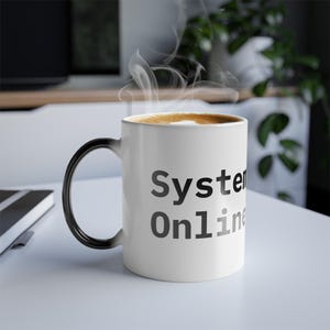 May include: A white ceramic mug with a black handle, filled with a hot drink. The mug displays the words "System Online" in grey lettering. Steam rises from the beverage, indicating it is freshly made.