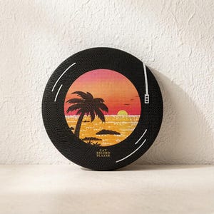 Puede incluir: Un rascador para gatos redondo y negro dise&ntilde;ado para parecer un disco de vinilo. El centro presenta una ilustraci&oacute;n de un atardecer con una palmera y olas. El texto CAT RECORD PLAYER est&aacute; impreso en dorado en la parte inferior de la superficie.
