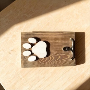 May include: A wooden bottle opener with a paw print design. The opener is made of dark brown wood and features a light-colored paw print. A silver-colored bottle opener is attached to the right side of the wooden board.