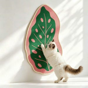 May include: A wall-mounted cat scratching pad shaped like a large leaf with a dark green center and pink border. The textured surface features cut-out leaf details and a light-colored piped edge. A fluffy white and brown cat is scratching the pad.