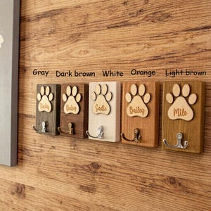 May include: A row of five wooden pet leash holders in varying colors: gray, dark brown, white, orange, and light brown. Each holder features a paw print design and a name, with a metal hook below.