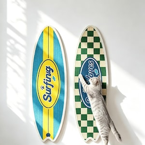 Puede incluir: Dos rascadores para gatos con forma de tabla de surf. El de la izquierda es azul y amarillo con el texto "Surfing" en un óvalo amarillo. El de la derecha presenta un patrón de cuadros verdes y blancos con un óvalo azul con el texto "Waves".