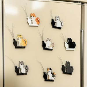 May include: A collection of cat-shaped magnets in various colors, including orange, white, black, and gray. Each magnet features a cat holding a black vase with a sprig of decorative elements. The magnets are attached to a beige refrigerator door.