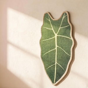 K&ouml;nnte beinhalten: Ein Wandbehang aus Fasern in Form eines gro&szlig;en, l&auml;nglichen Blattes. Das Blatt besteht aus dunkelgr&uuml;nem, gewebtem Material mit einer markanten, hellen Mittelrippe und verzweigten Seitenadern, eingefasst von einem dicken, cremefarbenen Rand.