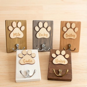May include: Five wooden pet leash holders with paw print designs and the names Milo, Rocky, Bailey, Sadie, and Daisy. Each holder has a metal hook. The wood tones vary from light to dark.