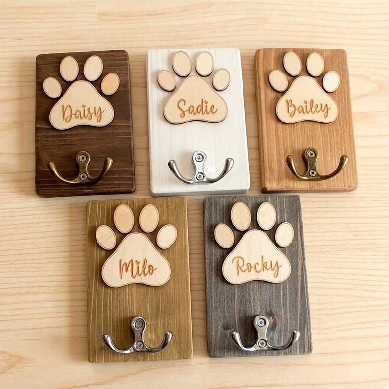 May include: Five wooden pet leash holders with paw print designs. Each holder has a different name: Daisy, Sadie, Bailey, Milo, and Rocky. The holders are in various colors, including brown, white, and gray, and feature metal hooks for hanging.
