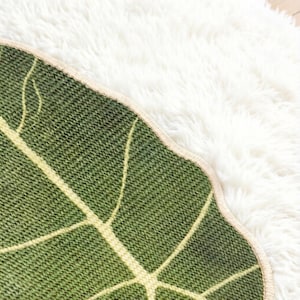 K&ouml;nnte beinhalten: Ein blattf&ouml;rmiger gr&uuml;ner Teppich mit einer strukturierten Strickoberfl&auml;che und hellgelben Blattadern. Der Teppich hat einen geschwungenen, gewellten Rand mit einer hellbeigen Einfassung und zeigt ein detailliertes botanisches Muster.
