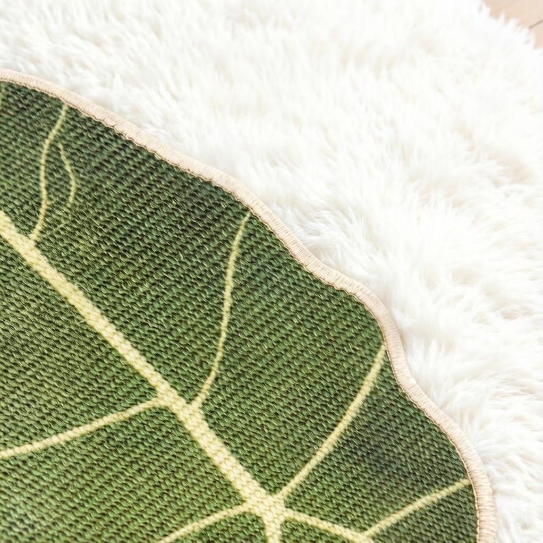 K&ouml;nnte beinhalten: Ein blattf&ouml;rmiger gr&uuml;ner Teppich mit einer strukturierten Strickoberfl&auml;che und hellgelben Blattadern. Der Teppich hat einen geschwungenen, gewellten Rand mit einer hellbeigen Einfassung und zeigt ein detailliertes botanisches Muster.