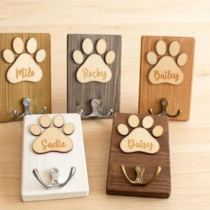 May include: Five wooden pet leash holders with paw print designs. Each holder has a different wood stain and a metal hook. The names "Milo", "Rocky", "Bailey", "Sadie", and "Daisy" are laser-cut on each paw print.