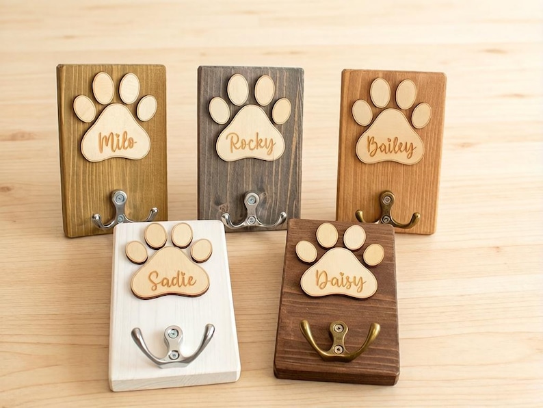 May include: Five wooden pet leash holders with paw print designs. Each holder has a different wood stain and a metal hook. The names "Milo", "Rocky", "Bailey", "Sadie", and "Daisy" are laser-cut on each paw print.