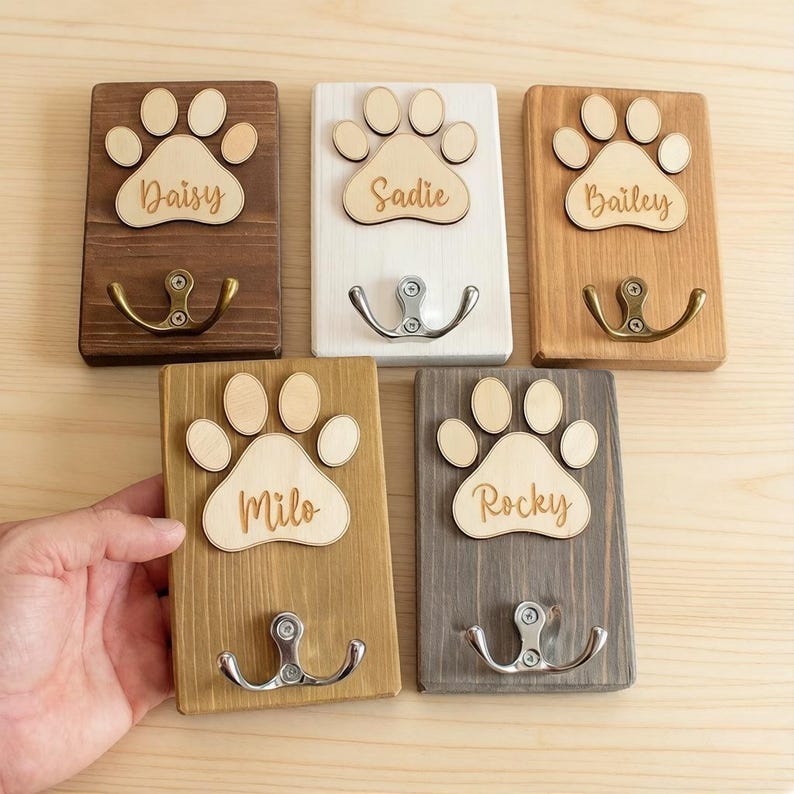 May include: Wooden pet leash holders with paw print designs. Each holder features a name: Daisy, Sadie, Bailey, Milo, and Rocky. The holders have metal hooks for leashes and are in various wood tones.