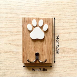 May include: A wooden wall hook with a white paw print design. The hook is 5.5 inches tall and 3.5 inches wide, featuring two silver hooks below the paw print. The wood has a natural grain.