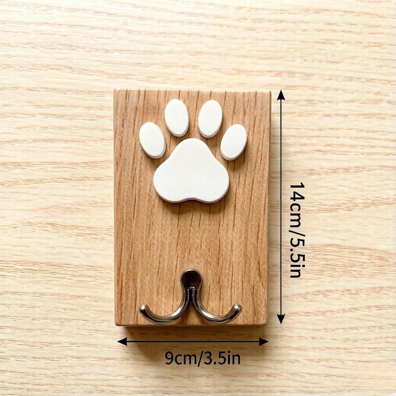 May include: A wooden wall hook with a white paw print design. The hook is 5.5 inches tall and 3.5 inches wide, featuring two silver hooks below the paw print. The wood has a natural grain.