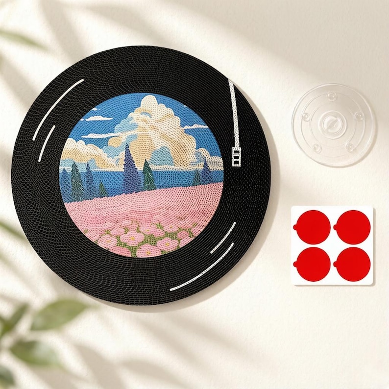 Puede incluir: Una decoraci&oacute;n de pared circular negra dise&ntilde;ada para parecer un disco de vinilo, con una ilustraci&oacute;n central de un paisaje con flores rosas, &aacute;rboles verdes y nubes azules. Incluye una ventosa transparente y cuatro c&iacute;rculos adhesivos rojos.