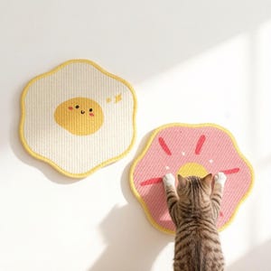 以下が含まれることがあります： 縁が波打った円形の壁掛けマット2枚。1枚はクリーム色で、中央に笑顔の黄色い目玉焼きのグラフィックが描かれています。もう1枚はピンク色で、中央が黄色く、赤い放射状の線が描かれています。