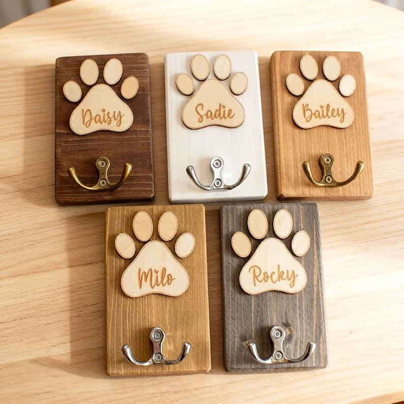 May include: A collection of personalized wooden wall hooks, each featuring a paw print design and a pet's name: Daisy, Sadie, Bailey, Milo, and Rocky. The hooks have different finishes, including brown, white, and gray, with metal hooks for hanging items.