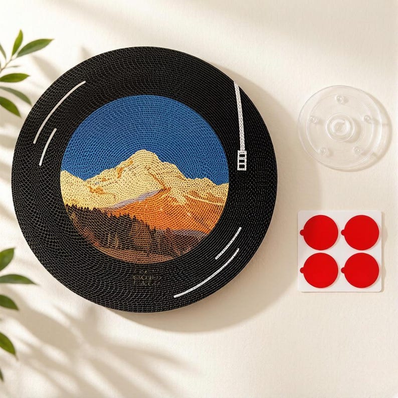 Puede incluir: Una pieza de arte mural circular dise&ntilde;ada para parecer un disco de vinilo, con una imagen central estilo mosaico de un pico de monta&ntilde;a dorado bajo un cielo azul, acompa&ntilde;ada de una ventosa transparente y cuatro c&iacute;rculos adhesivos rojos.