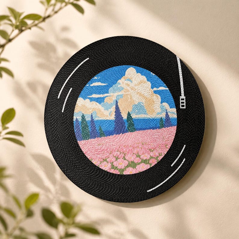 Puede incluir: Una pieza de arte mural circular dise&ntilde;ada para parecer un disco de vinilo negro. El centro presenta una escena de paisaje bordada con un campo de flores rosas, &aacute;rboles oscuros, un horizonte azul y grandes nubes blancas.