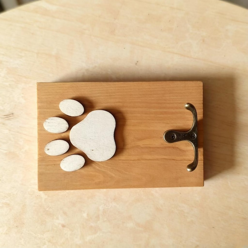 May include: A wooden wall hook with a paw print design. The rectangular board is light brown with a white paw print and a bronze-colored hook. The paw print is made of five white oval shapes and one larger white shape.