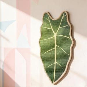 K&ouml;nnte beinhalten: Ein Wandbehang in Form eines gro&szlig;en Blattes mit einer gewebten Textur in dunklen und hellen Gr&uuml;nt&ouml;nen. Das Blatt weist eine markante Mittelrippe und hellgelbe Blattadern auf und ist mit einem dicken, hellfarbenen Rand versehen.