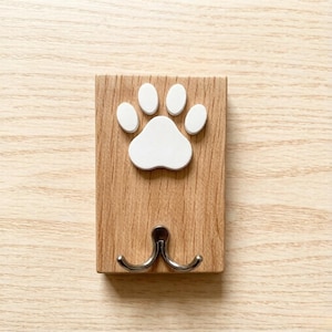 May include: A wooden wall hook with a white paw print design. The hook has two silver-colored prongs for hanging items. The wood has a natural grain pattern.
