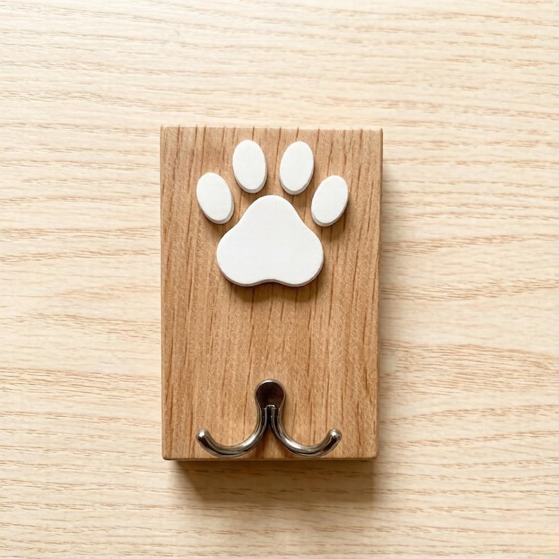 May include: A wooden wall hook with a white paw print design. The hook has two silver-colored prongs for hanging items. The wood has a natural grain pattern.