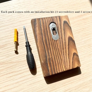 May include: A wooden wall hanging with a metal bracket, a black screwdriver, a yellow wall anchor, and a screw. The text "Each pack comes with an installation kit (1 screwdriver and 1 screw)" is visible.