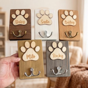 May include: Five wooden wall hooks with paw print designs and personalized names. The hooks are in various colors: dark brown, white, light brown, gold, and gray. Each hook has a metal hook for hanging items.