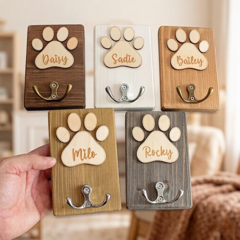 May include: Five wooden wall hooks with paw print designs and personalized names. The hooks are in various colors: dark brown, white, light brown, gold, and gray. Each hook has a metal hook for hanging items.