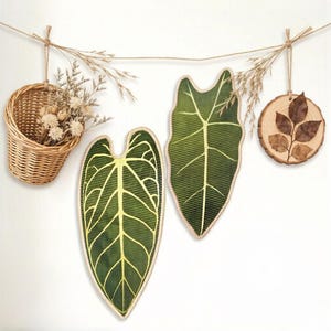 K&ouml;nnte beinhalten: Ein Wandbehang-Set bestehend aus einem kleinen geflochtenen Weidenkorb mit Trockenblumen, zwei stoffartigen blattf&ouml;rmigen Wanddekorationen mit dunkelgr&uuml;nem und gelbem Adernmuster sowie einem runden Holzscheiben-Ornament mit braunem Blatt.