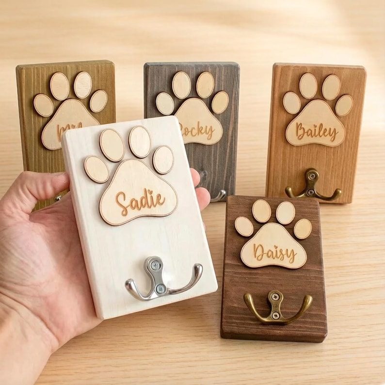 May include: Wooden pet leash hooks in various colors, each featuring a paw print design and a name. The hooks are made of wood and have a metal hook for hanging leashes or other items. The names include "Sadie", "Rocky", "Bailey", "Daisy", and "Mia".