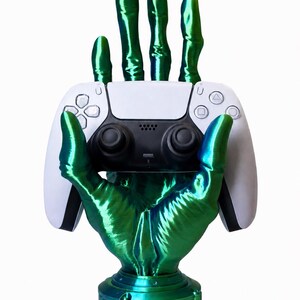 May include: A metallic green, multi-fingered hand sculpture mounted on a circular base, holding a white and black wireless video game controller. The hand features a glossy, iridescent finish with visible 3D printed texture lines throughout.