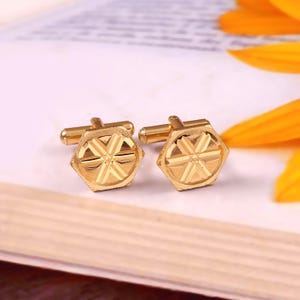 Puede incluir: Un par de gemelos dorados de forma hexagonal con un patrón central de estrella de seis puntas. Cada gemelo tiene un acabado pulido con detalles texturizados en la superficie y un mecanismo de cierre giratorio estándar en la parte posterior.