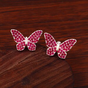 Puede incluir: Un par de pendientes de botón con forma de mariposa que presentan un marco metálico de tono plateado incrustado con numerosas piedras circulares pequeñas de color rosa vibrante que cubren toda la superficie de las alas.