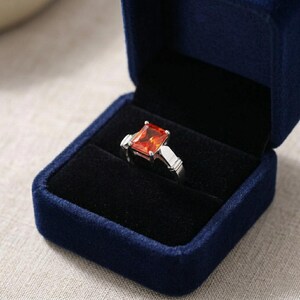 May include: A silver-toned ring featuring a rectangular, faceted orange gemstone set in a four-prong mount, displayed inside a dark blue velvet jewelry box with a black interior lining that holds the ring securely in place for presentation.