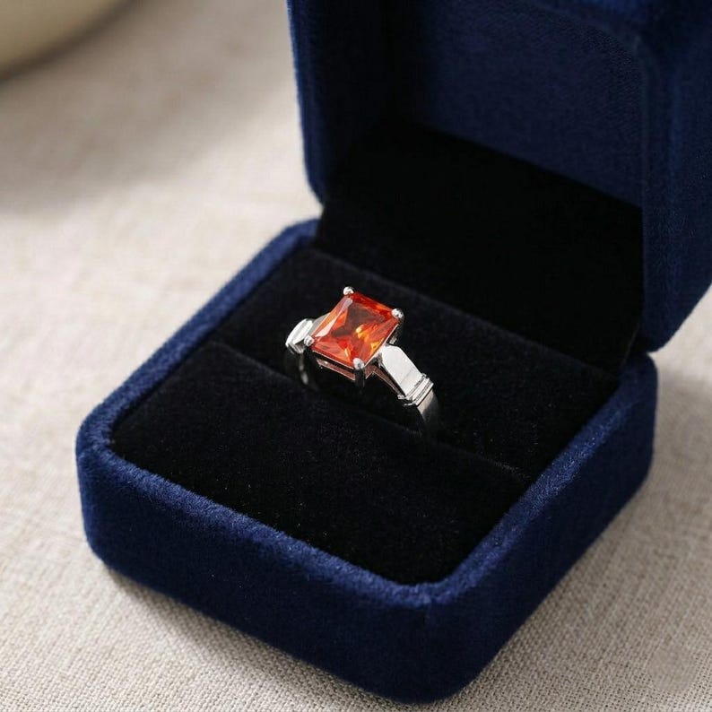 May include: A silver-toned ring featuring a rectangular, faceted orange gemstone set in a four-prong mount, displayed inside a dark blue velvet jewelry box with a black interior lining that holds the ring securely in place for presentation.