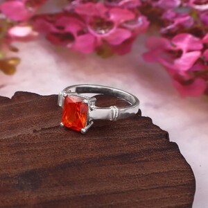 May include: A silver-toned ring featuring a rectangular, vibrant orange gemstone set in a four-prong mount. The band has a smooth, polished finish with a subtle decorative grooved detail on each side near the central stone setting.