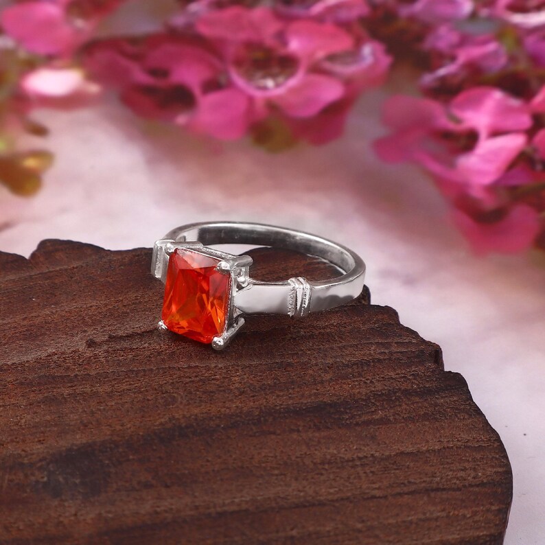 May include: A silver-toned ring featuring a rectangular, vibrant orange gemstone set in a four-prong mount. The band has a smooth, polished finish with a subtle decorative grooved detail on each side near the central stone setting.