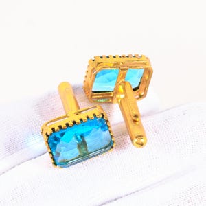 Puede incluir: Un par de gemelos dorados con centros de piedras preciosas rectangulares de color azul claro. Los gemelos tienen un borde dorado detallado y texturizado y un cierre de poste clásico. Las piedras preciosas son de un azul vibrante y transparente, engastadas en un marco dorado.