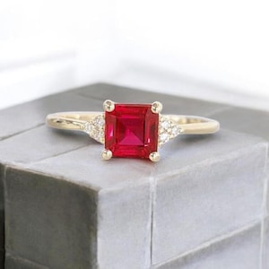 May include: A gold ring with a striking, square-cut, red gemstone at its centre. Small, clear stones are set on either side of the main stone. The ring is presented on a grey surface, highlighting its elegant design and craftsmanship.