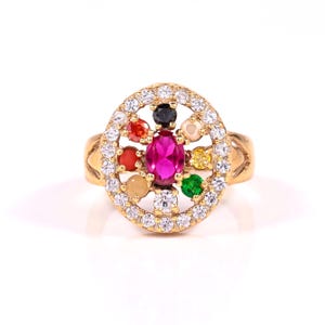 May include: A gold ring featuring a central oval-cut pink gemstone surrounded by a halo of white gemstones. The ring's design includes a circular pattern with various colored gemstones, including red, green, yellow, and black, set within the circle.