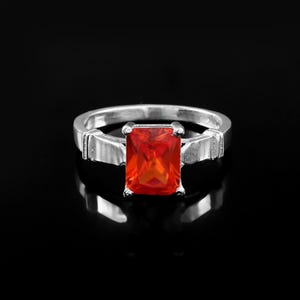 May include: A silver-toned ring featuring a rectangular, faceted orange gemstone set in a four-prong mount. The band has a smooth, polished finish with decorative metallic segments on either side of the central stone, creating a structured look.