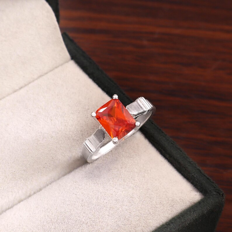 May include: A silver-toned ring featuring a rectangular, faceted orange gemstone set in a four-prong setting. The band has a smooth, polished finish with a slightly wider design near the stone, presented in a neutral-colored jewelry box.