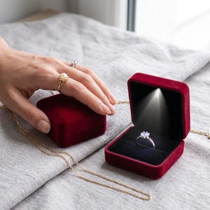 Op de afbeelding: Een vierkant, donkerrood fluwelen ringdoosje met een ingebouwd LED-lampje dat een zilveren ring met een heldere steen verlicht. Een tweede gesloten rood fluwelen doosje staat ernaast en een gouden ketting ligt op de ondergrond. De binnenkant is zwart.